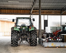 A large green tractor and other farming supplies sit in a large warehouse with an open garage door.