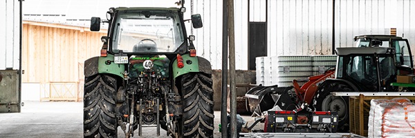 A large green tractor and other farming supplies sit in a large warehouse with an open garage door.
