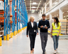 Three adults walk through a warehouse of goods and peer at a tablet one of them is holding.