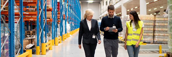 Three adults walk through a warehouse of goods and peer at a tablet one of them is holding.