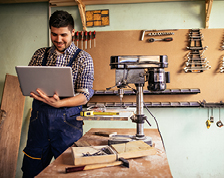 A carpenter stands in his workshop wearing blue overalls and using a laptop. 