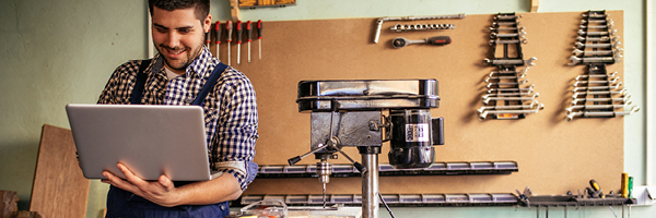 A carpenter stands in his workshop wearing blue overalls and using a laptop.