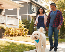 A couple with a large white, fluffy dog take a walk outside a white home with a large front porch.