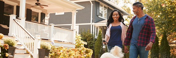 A couple with a large white, fluffy dog take a walk outside a white home with a large front porch.