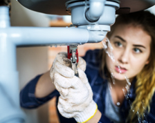 Woman uses tools to fix plumbing under a sink.