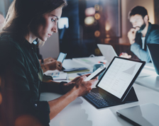 A businesswoman sits at a table with her peers comparing data on her smartphone and tablet.