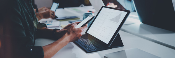 A businesswoman sits at a table with her peers comparing data on her smartphone and tablet.