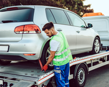 A man in a neon safety vest fastens the rear wheel of a silver car to a flatbed tow truck.