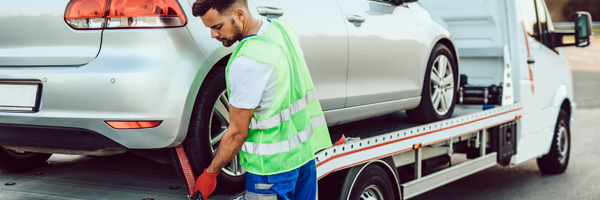 A man in a neon safety vest fastens the rear wheel of a silver car to a flatbed tow truck.