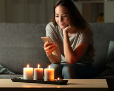 A woman sitting on a gray couch holding a smartphone sits near a table with three lit candles.
