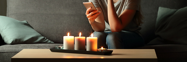 A woman sitting on a gray couch holding a smartphone sits near a table with three lit candles.