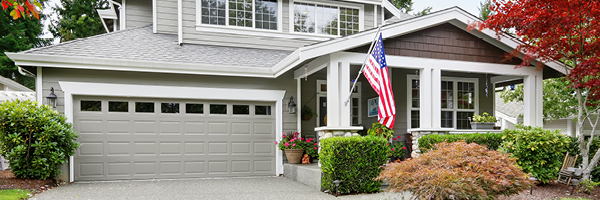 A grey two-story home with white window trim and an American flag hanging off the front porch.