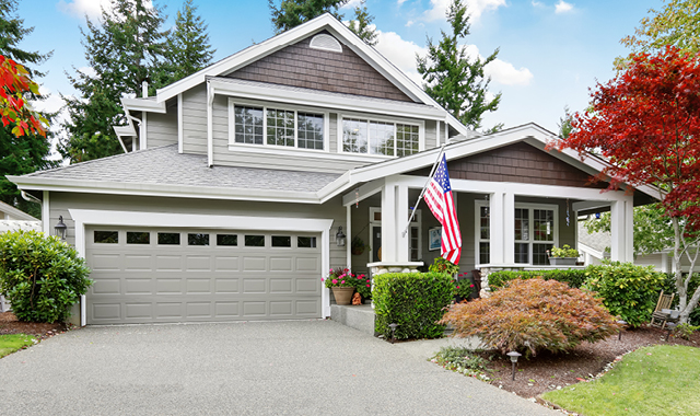 A grey two-story home with white window trim and an American flag hanging off the front porch.