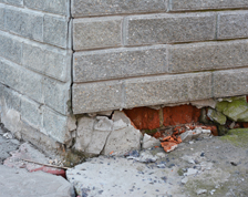 A gray brick exterior of a building crumbles where the foundation meets the ground.