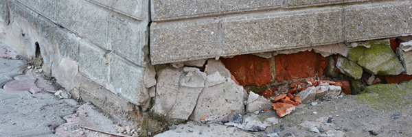 A gray brick exterior of a building crumbles where the foundation meets the ground.