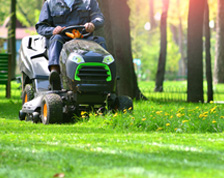 A lawn care contractor drives a riding lawn mower around a park with trees and benches.