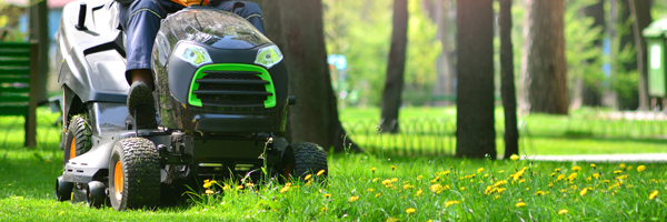 A lawn care contractor drives a riding lawn mower around a park with trees and benches.