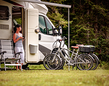 A woman in a striped tank top and white pants holds a mug while standing outside her parked RV.