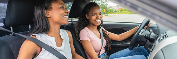 A teenage girl and her mother sit in the front seats of a vehicle with their seatbelts on.