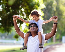 A man carries a young girl on his shoulders outside. They hold hands and smile at each other. 