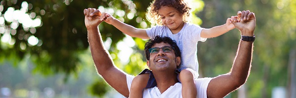 A man carries a young girl on his shoulders outside. They hold hands and smile at each other. 
