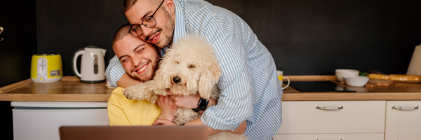 Two men with a puppy wrap their arms around each other while looking at a laptop and smiling.