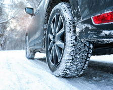 Two tires on a dark vehicle roll over a road that is covered in snow.